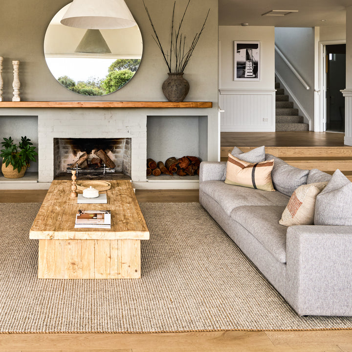 A beige wool and jute rug placed on a wooden floor under a grey sofa, with a white fireplace and round mirror in the background.