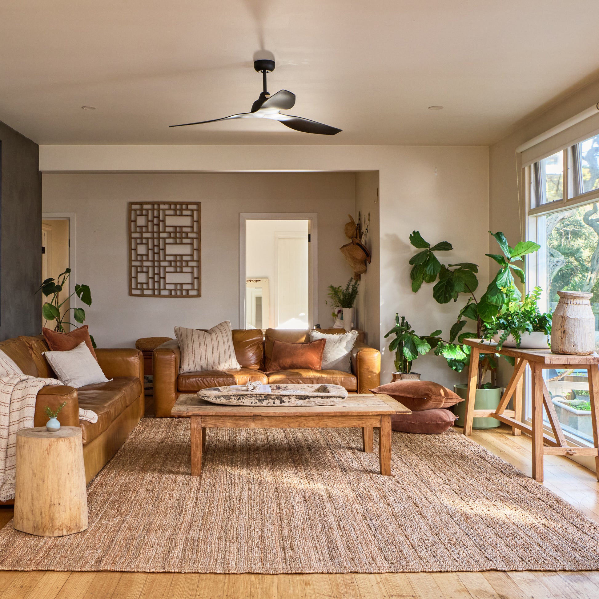 A styled living room interior featuring a brown leather sofa, a wooden coffee table, a large natural jute area rug, a ceiling fan, and multiple potted green plants near a bright window.