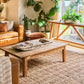 A rustic wooden coffee table in a warm, modern boho living room setting with a large woven jute rug, leather sofa, and potted plants.