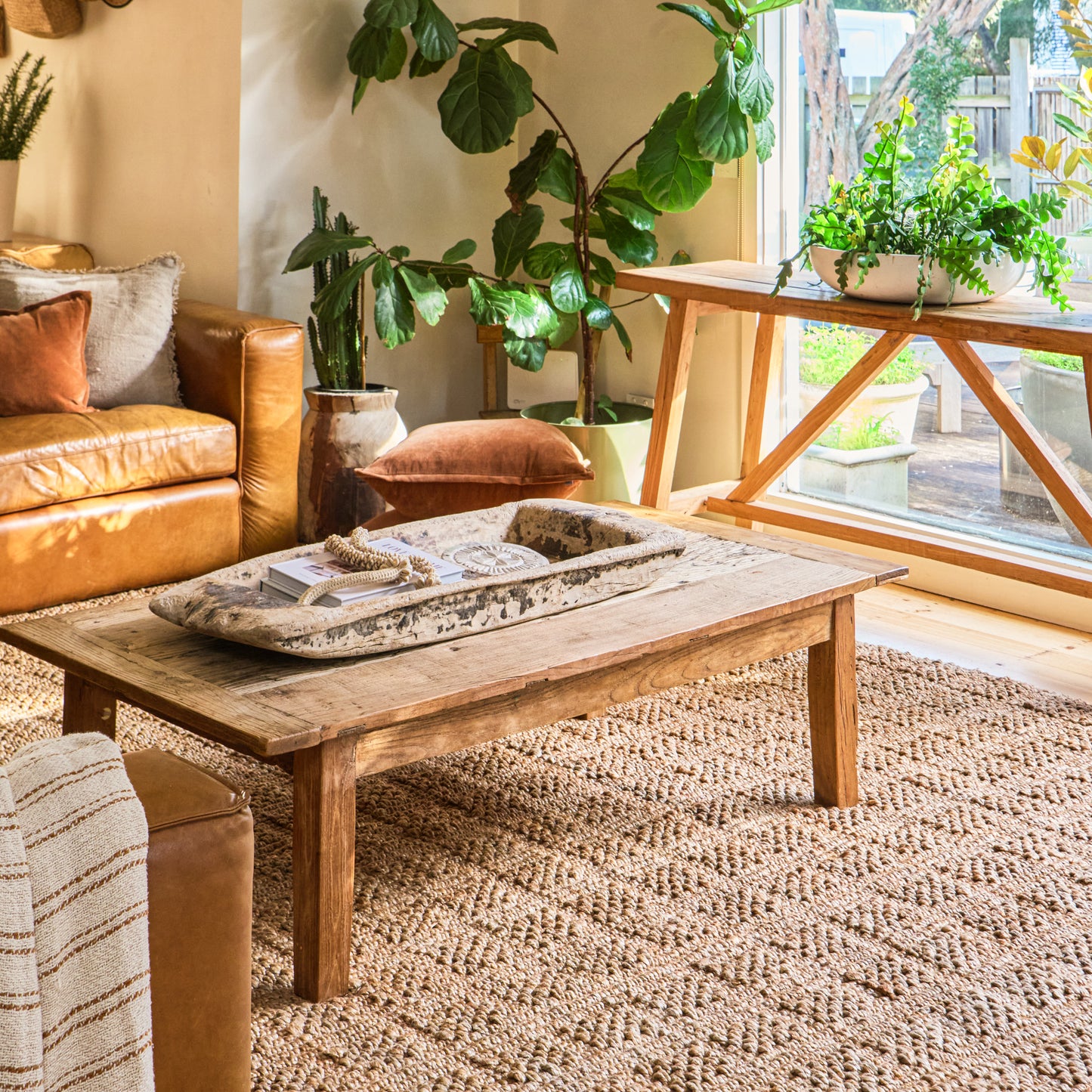 A rustic wooden coffee table in a warm, modern boho living room setting with a large woven jute rug, leather sofa, and potted plants.