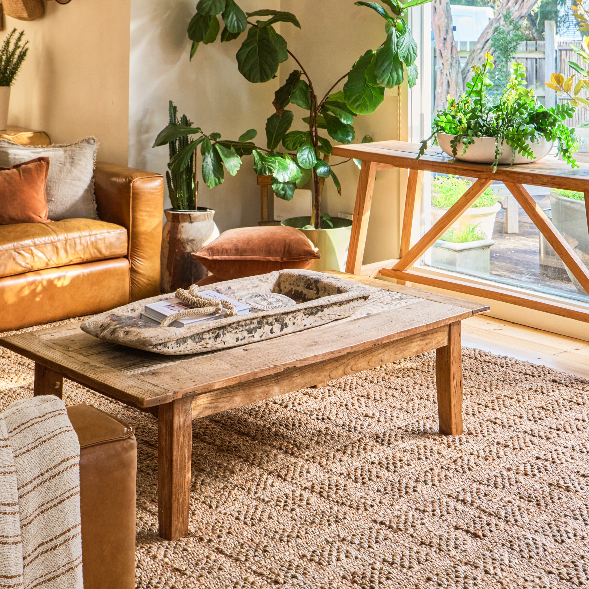 A rustic wooden coffee table in a warm, modern boho living room setting with a large woven jute rug, leather sofa, and potted plants.