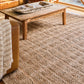 A textured natural jute area rug in warm beige and brown tones, shown in a cozy living room setting under a wooden coffee table and next to a leather sofa with a striped throw blanket.