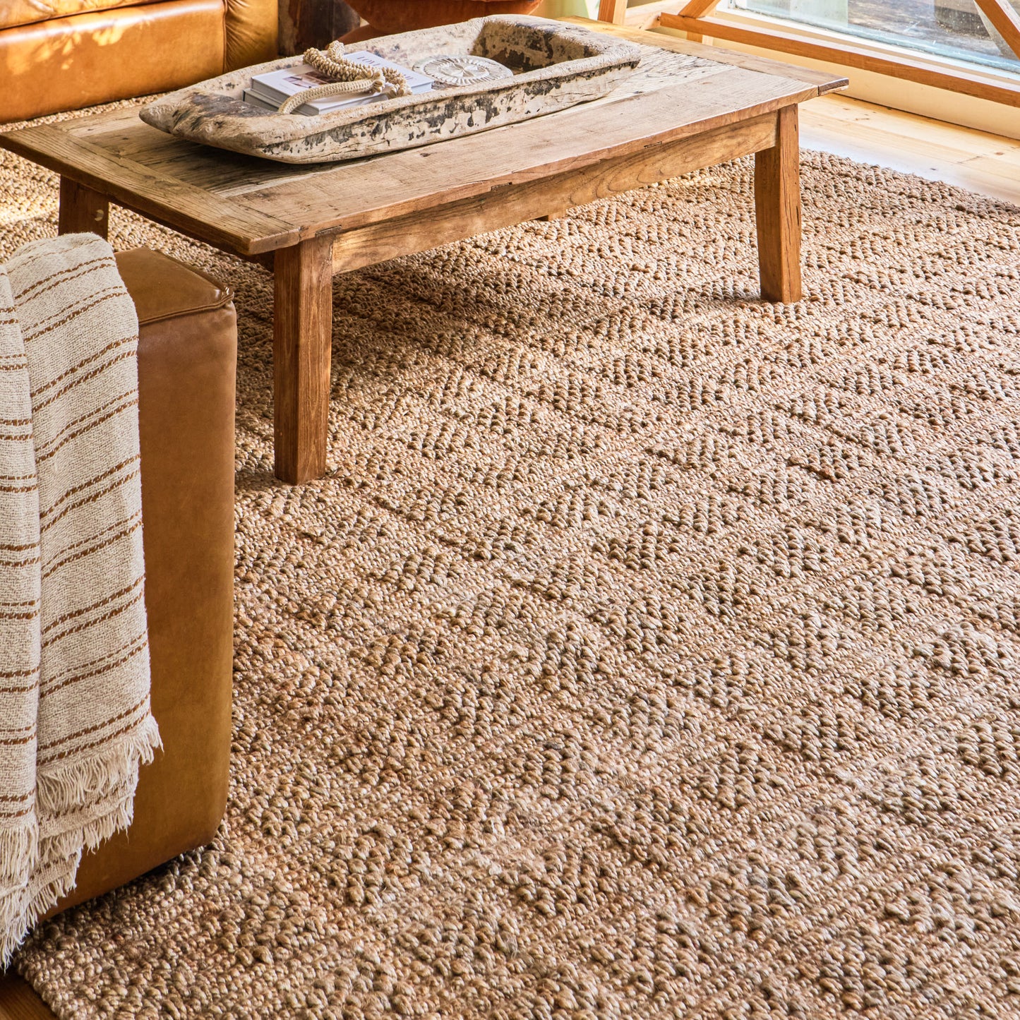 A textured natural jute area rug in warm beige and brown tones, shown in a cozy living room setting under a wooden coffee table and next to a leather sofa with a striped throw blanket.