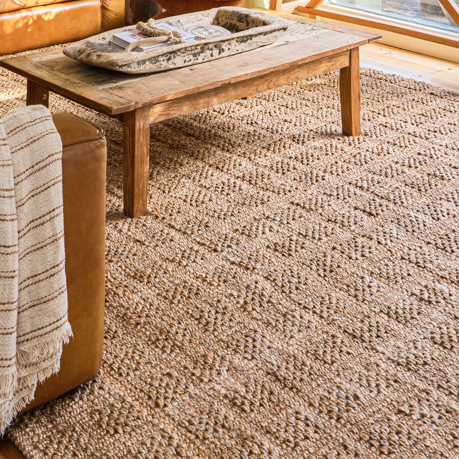 A textured natural jute area rug in warm beige and brown tones, shown in a cozy living room setting under a wooden coffee table and next to a leather sofa with a striped throw blanket.