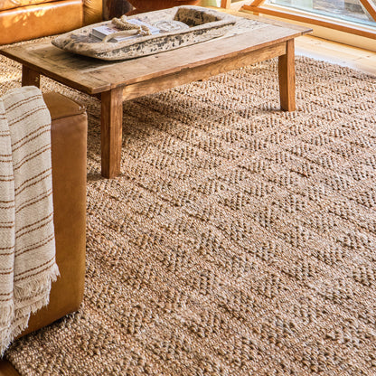 A textured natural jute area rug in warm beige and brown tones, shown in a cozy living room setting under a wooden coffee table and next to a leather sofa with a striped throw blanket.