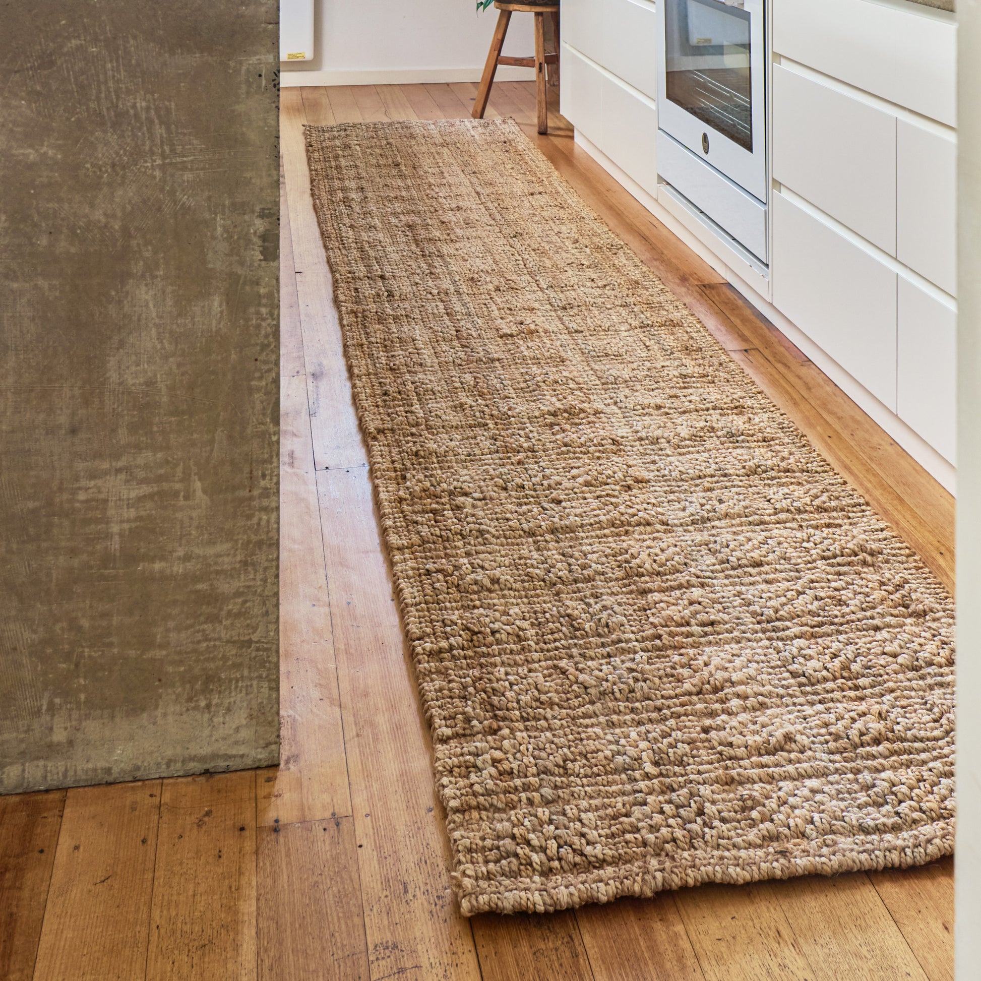 A long, low-pile natural jute or sisal runner rug in a warm beige color, laid out on a light hardwood floor in a modern kitchen setting.