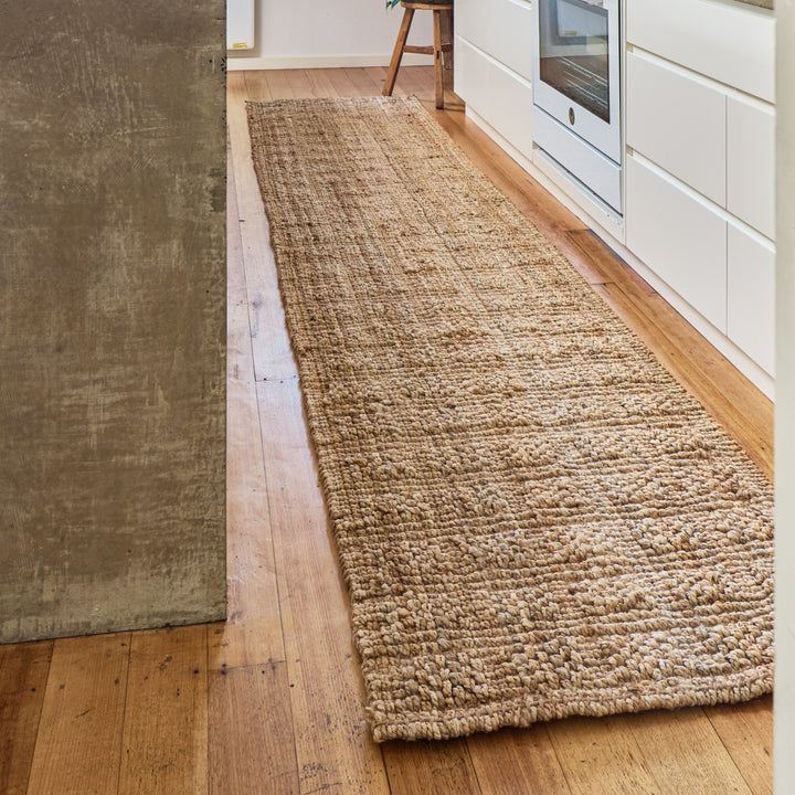 A long, low-pile natural jute or sisal runner rug in a warm beige color, laid out on a light hardwood floor in a modern kitchen setting.