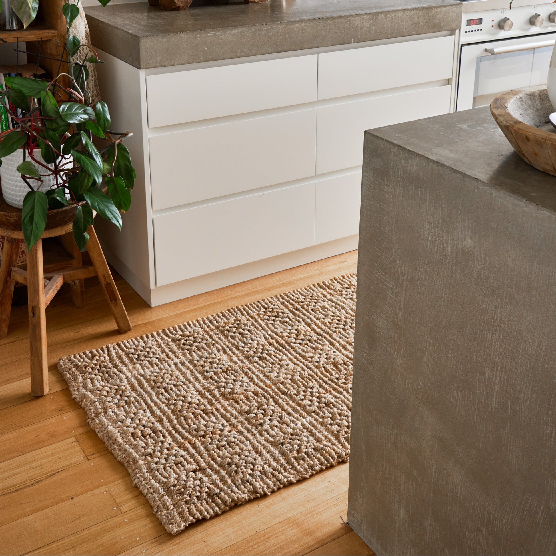 A styled kitchen interior featuring a textured beige woven area rug on a light wood floor in front of white cabinets and a concrete countertop.