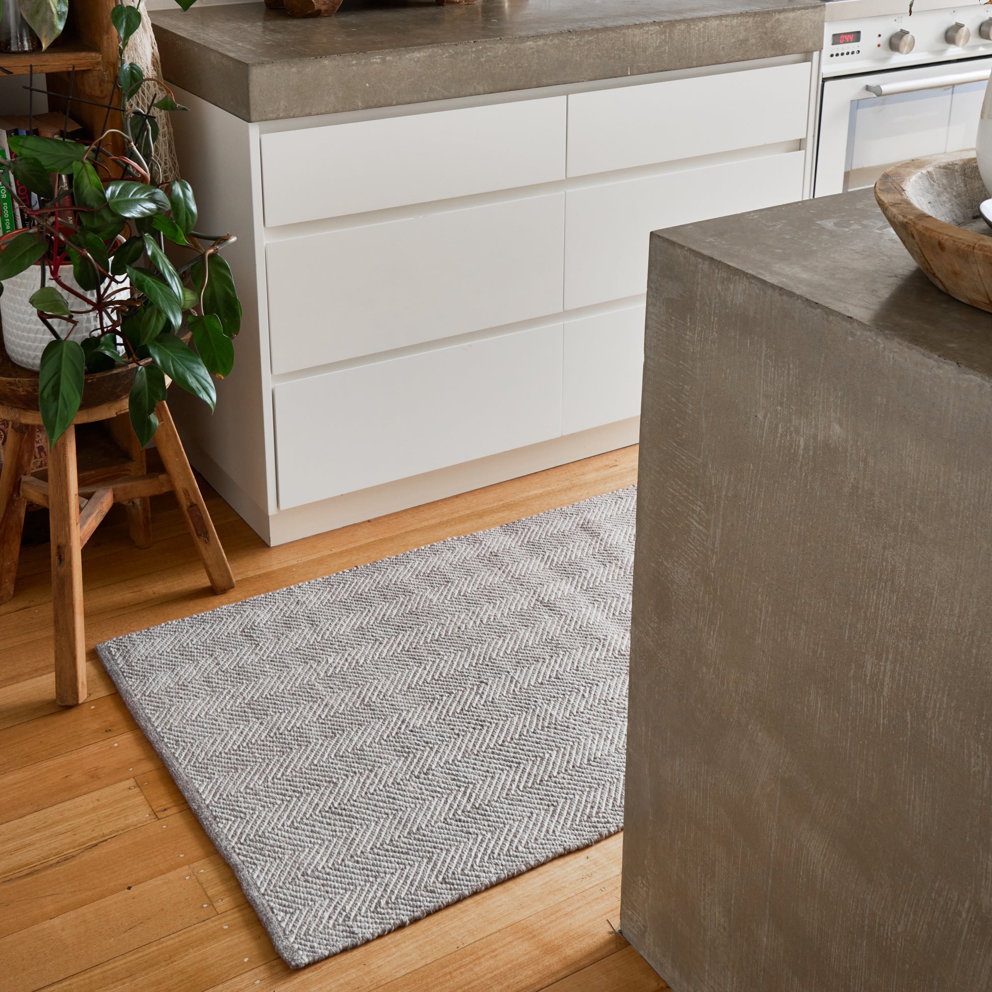 A gray and white herringbone patterned area rug on a light wood floor in a modern kitchen setting with white cabinets and concrete countertops.