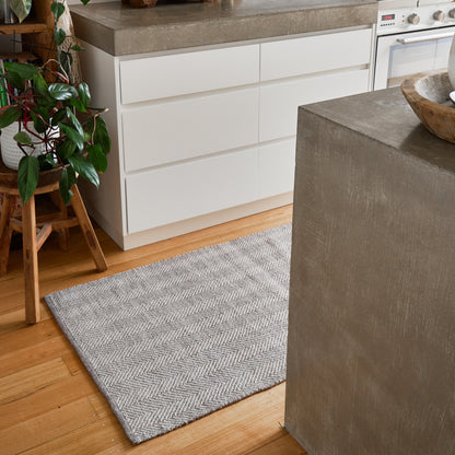 A gray and white herringbone patterned area rug on a light wood floor in a modern kitchen setting with white cabinets and concrete countertops.