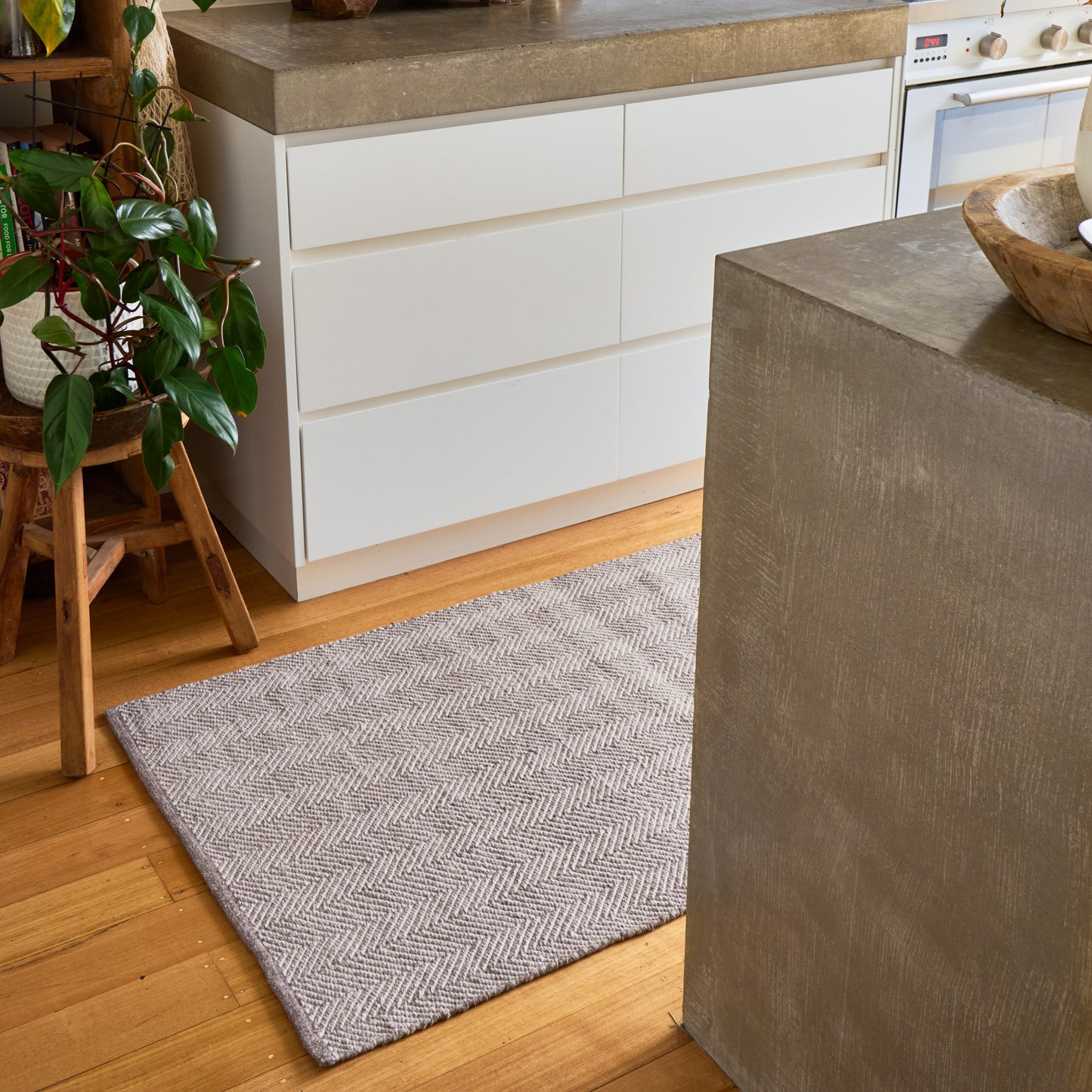A rectangular area rug with a gray and white herringbone pattern, placed on a light wood floor in a modern kitchen setting.