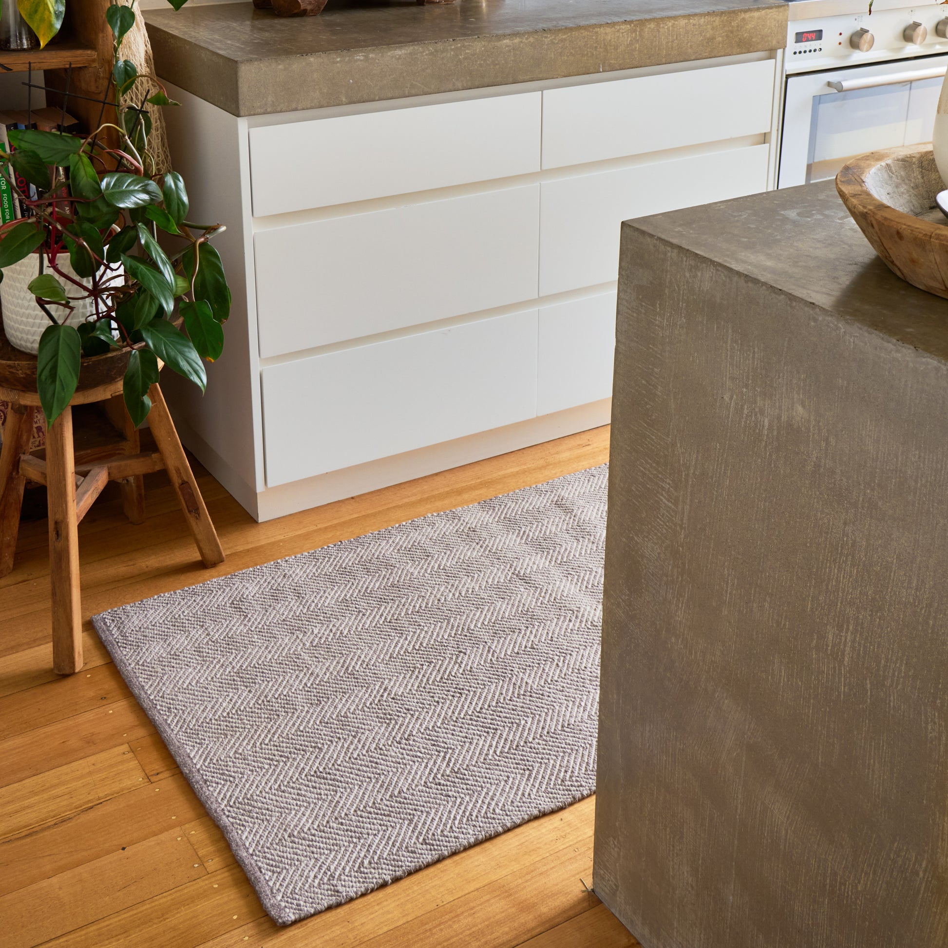A rectangular area rug with a gray and white herringbone pattern, placed on a light wood floor in a modern kitchen setting.
