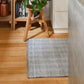 A gray and white herringbone-patterned rectangular rug on a light wood floor in a home setting, with a wooden stool, books, and a plant in the background.