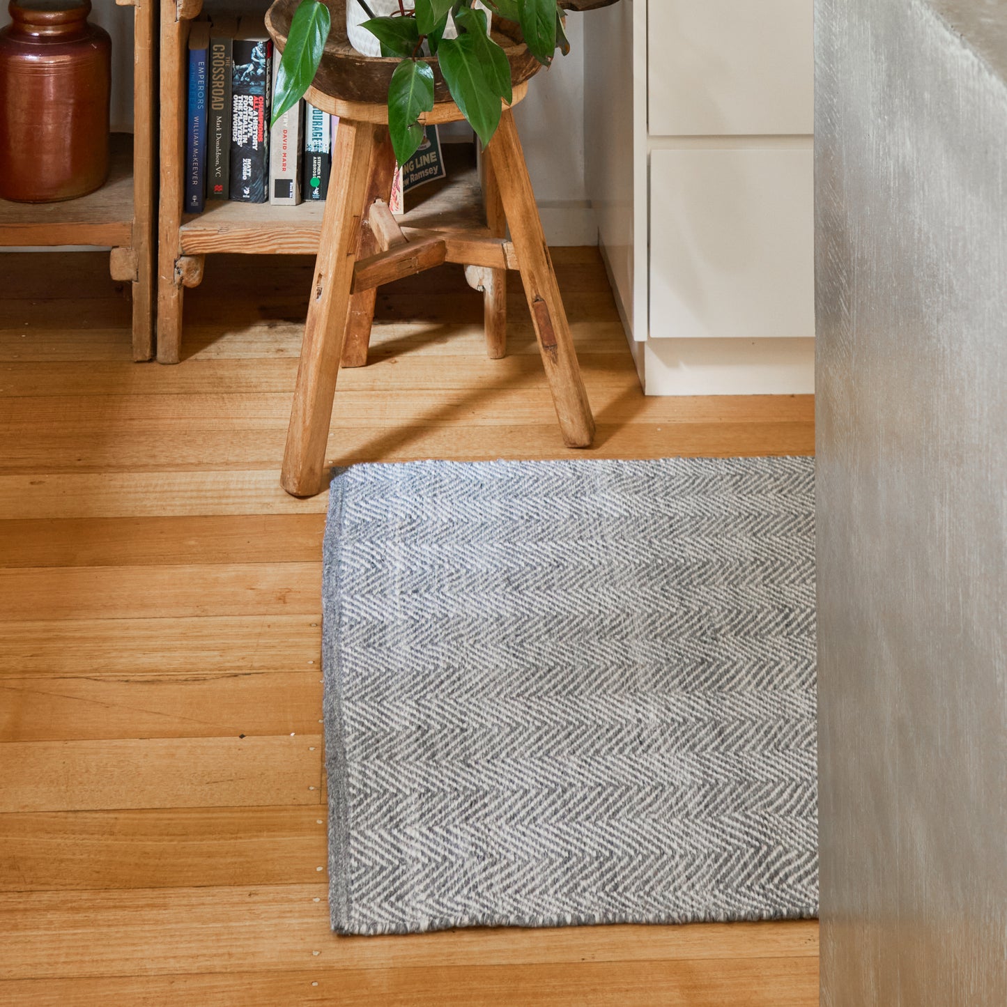 A gray and white herringbone-patterned rectangular rug on a light wood floor in a home setting, with a wooden stool, books, and a plant in the background.