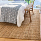 A natural jute area rug in a bedroom setting, shown under a bed with white linens and a blue patterned throw, with a rustic wooden bench and window in the background.