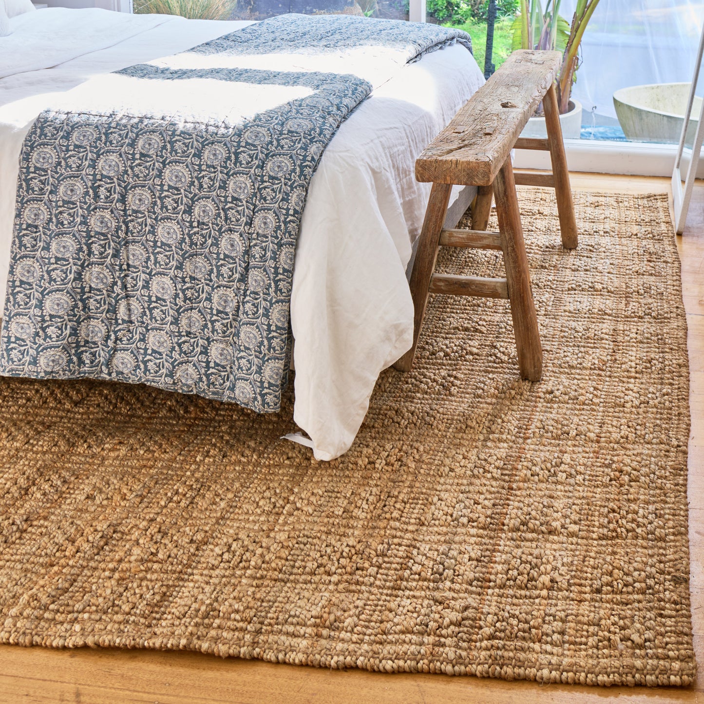A natural jute area rug in a bedroom setting, shown under a bed with white linens and a blue patterned throw, with a rustic wooden bench and window in the background.