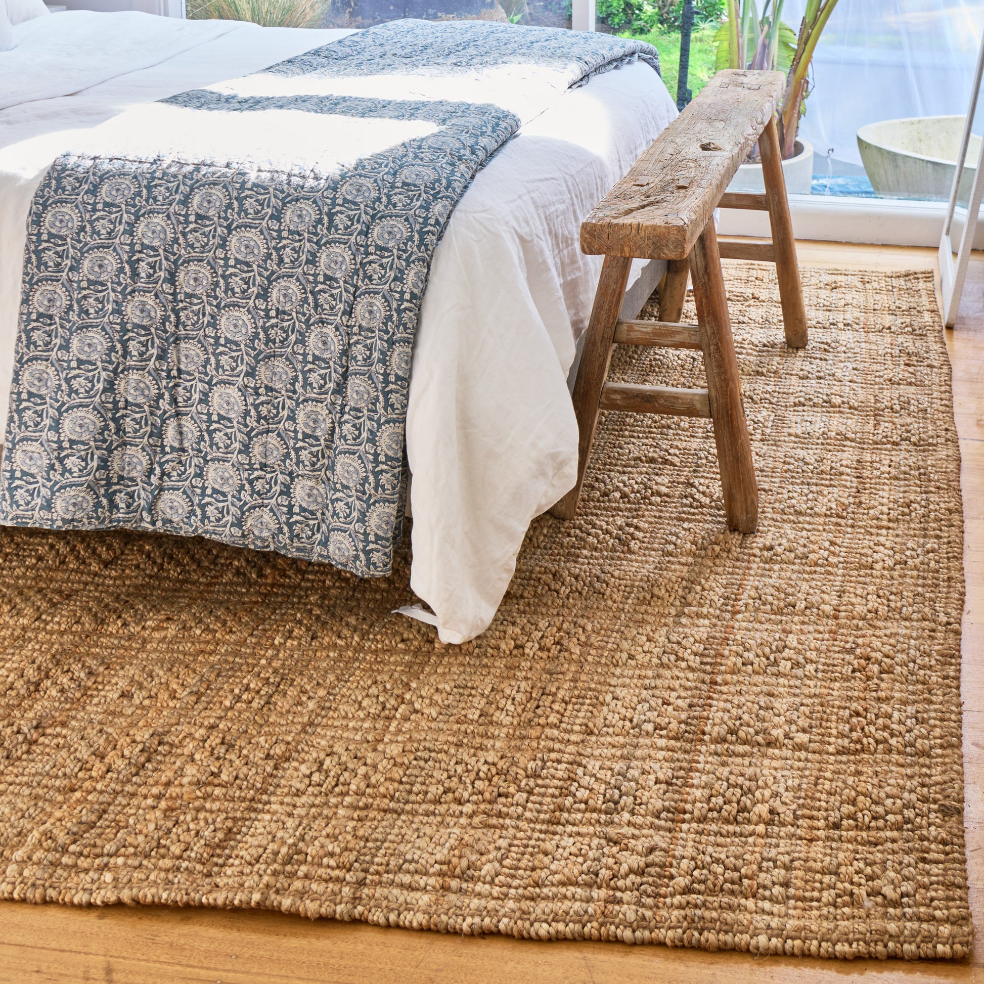 A natural jute area rug in a bedroom setting, shown under a bed with white linens and a blue patterned throw, with a rustic wooden bench and window in the background.