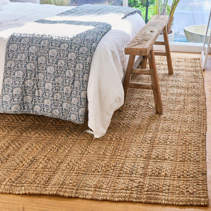A natural jute area rug in a bedroom setting, shown under a bed with white linens and a blue patterned throw, with a rustic wooden bench and window in the background.