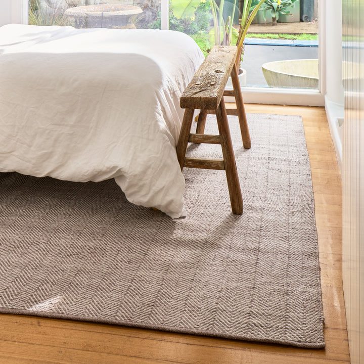 A rectangular woven area rug in a warm taupe-gray and beige herringbone pattern, styled in a bright bedroom next to a bed with white bedding and a rustic wooden stool.