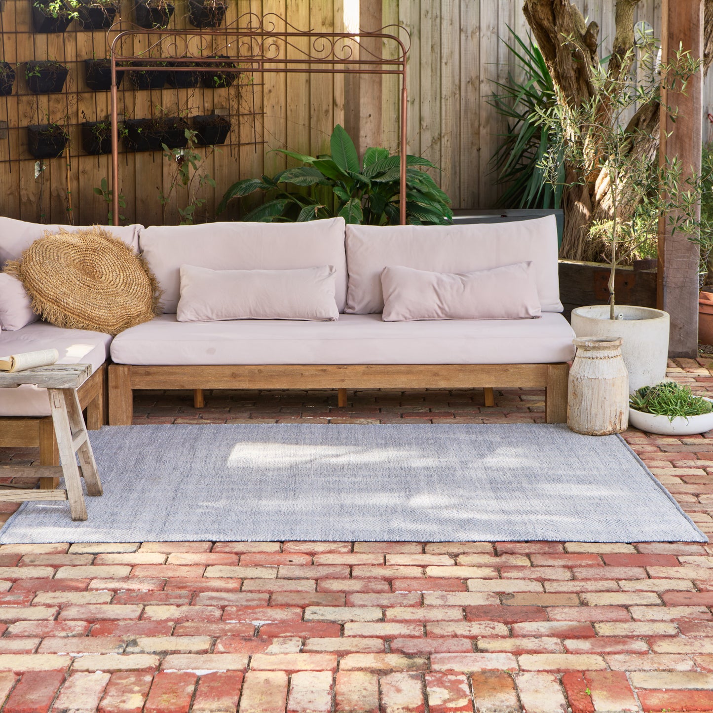 A light gray outdoor area rug laid on a brick patio in front of a wooden outdoor sofa with pale blush cushions, styled with a small woven stool, round straw pillow, and potted plants against a wooden fence.