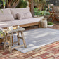 A light gray and white textured woven outdoor area rug on a brick patio, styled with a wooden bench, a rustic wooden stool, and potted plants.