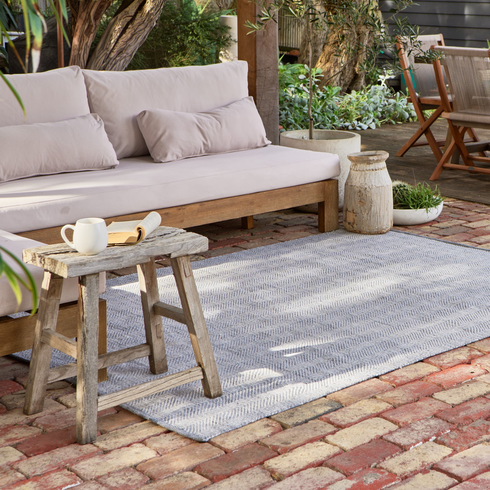 A light gray and white textured woven outdoor area rug on a brick patio, styled with a wooden bench, a rustic wooden stool, and potted plants.