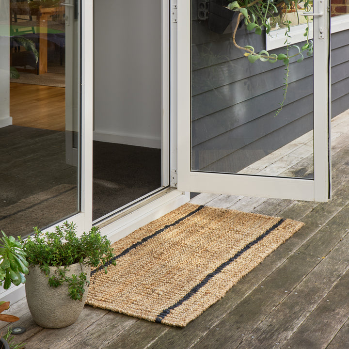 A rectangular natural jute doormat with two dark navy stripes, placed on a weathered wooden deck in front of an open white-framed glass door.