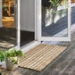 A rectangular woven jute doormat with natural tan and light brown stripes, placed on a dark wooden deck in front of an open white-framed glass door. Green potted plants are visible on the deck and patio.