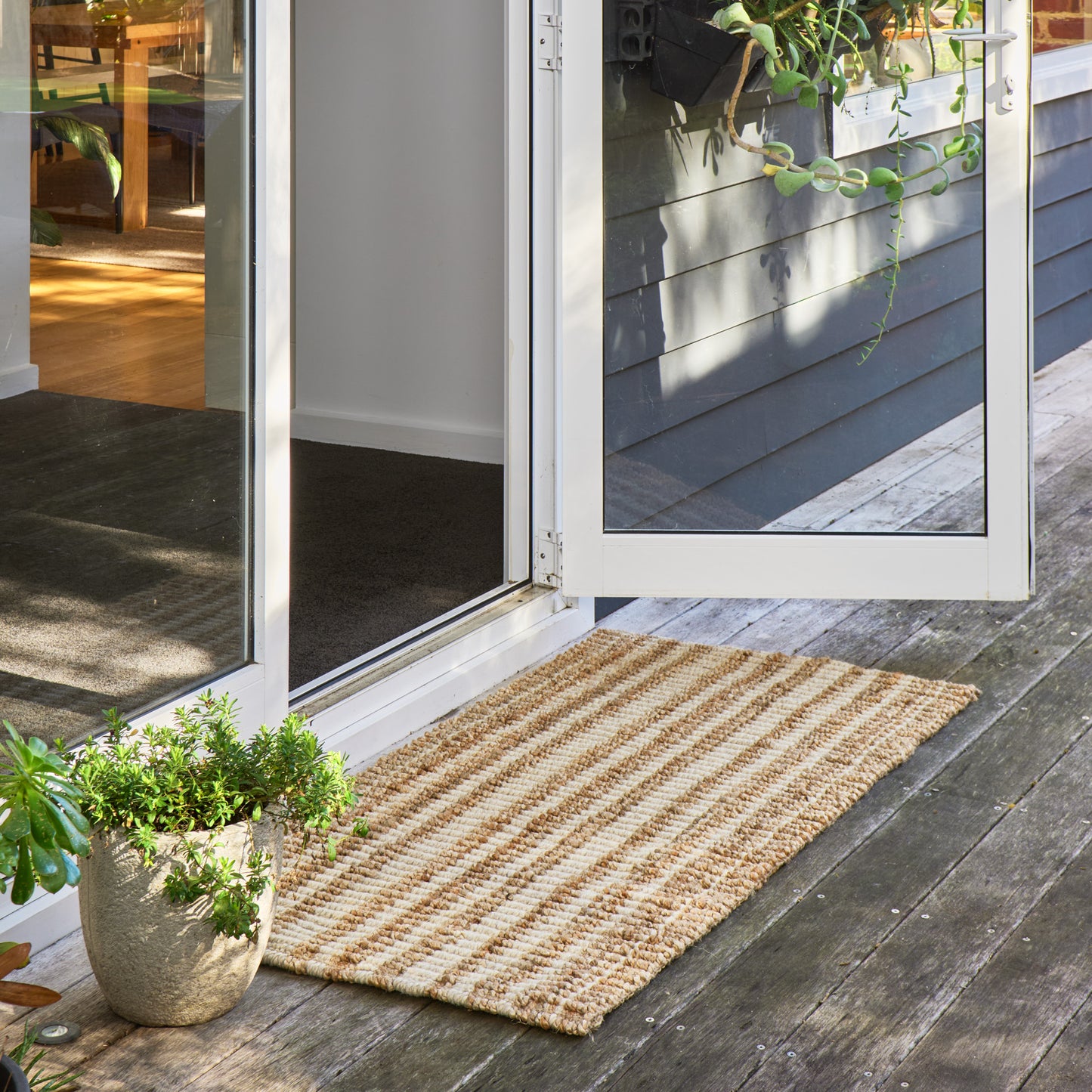 A rectangular woven jute doormat with natural tan and light brown stripes, placed on a dark wooden deck in front of an open white-framed glass door. Green potted plants are visible on the deck and patio.