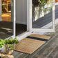 A rectangular woven jute doormat with a black horizontal stripe, placed on a weathered wooden deck in front of white-framed glass patio doors.