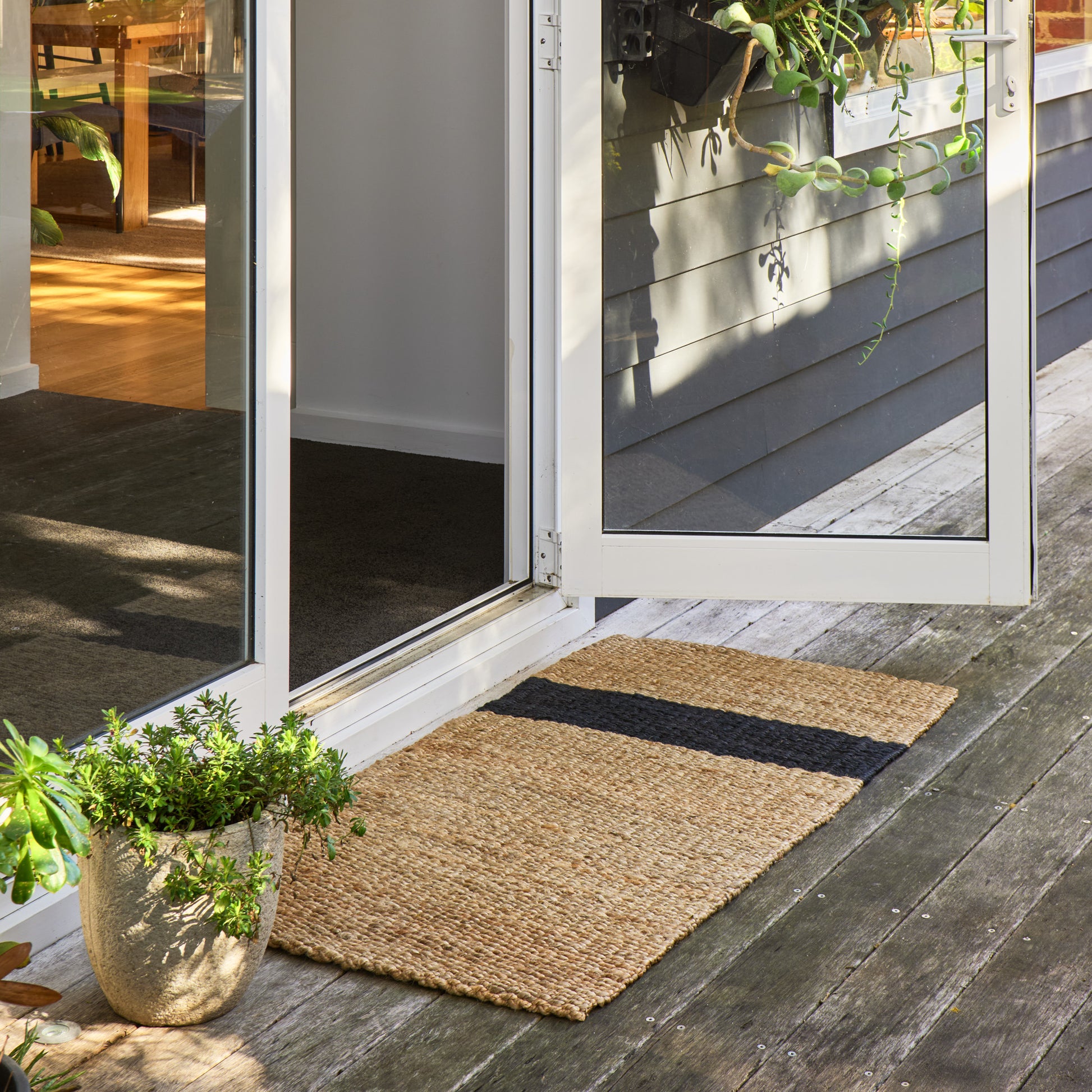 A rectangular woven jute doormat with a black horizontal stripe, placed on a weathered wooden deck in front of white-framed glass patio doors.