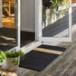 A rectangular black coir doormat with a horizontal natural beige stripe, placed on a weathered wooden deck in front of white-framed glass patio doors.