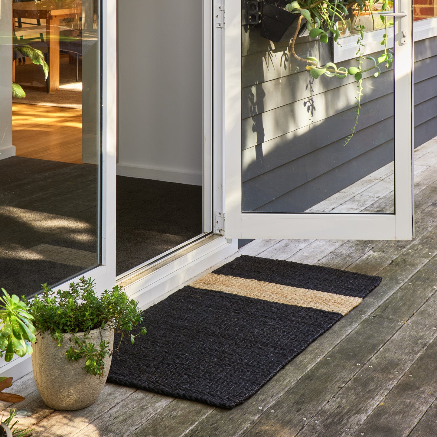 A rectangular black coir doormat with a horizontal natural beige stripe, placed on a weathered wooden deck in front of white-framed glass patio doors.