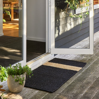 A rectangular black coir doormat with a horizontal natural beige stripe, placed on a weathered wooden deck in front of white-framed glass patio doors.