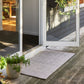 A rectangular light gray woven doormat with a subtle herringbone pattern, placed on a weathered wooden deck in front of an open white-framed glass patio door. Potted green plants are visible on the deck and reflected in the glass.
