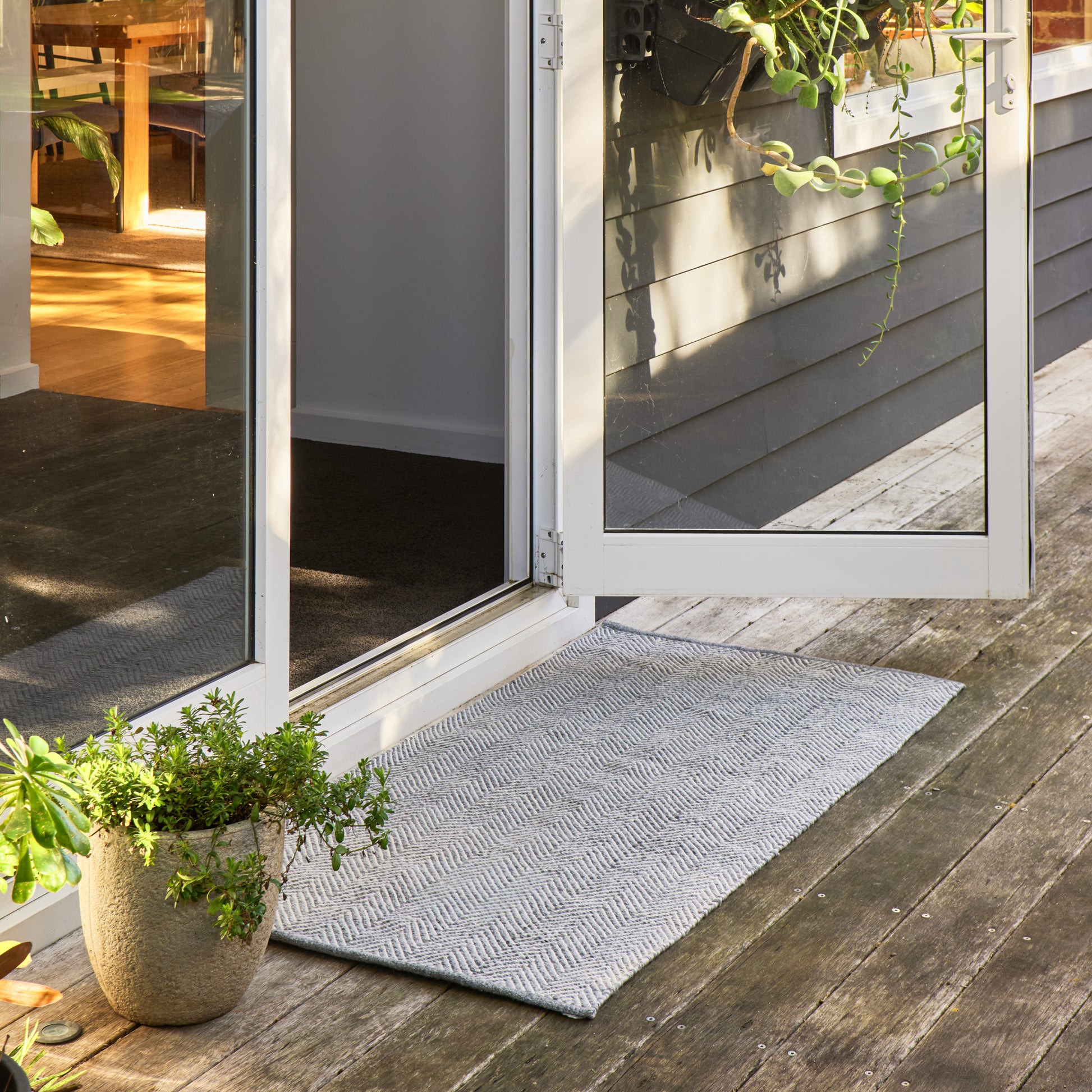 A light gray textured doormat with a subtle chevron pattern, positioned on a weathered wooden deck in front of white-framed glass patio doors.