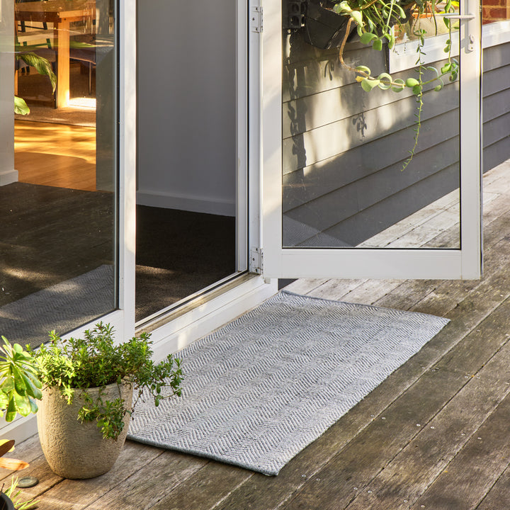 A light gray textured doormat with a subtle chevron pattern, positioned on a weathered wooden deck in front of white-framed glass patio doors.