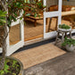 A natural woven jute or sisal doormat leading from a wooden deck onto a patio door into a furnished living room. The scene includes potted green plants and a rustic wooden bench on the patio.
