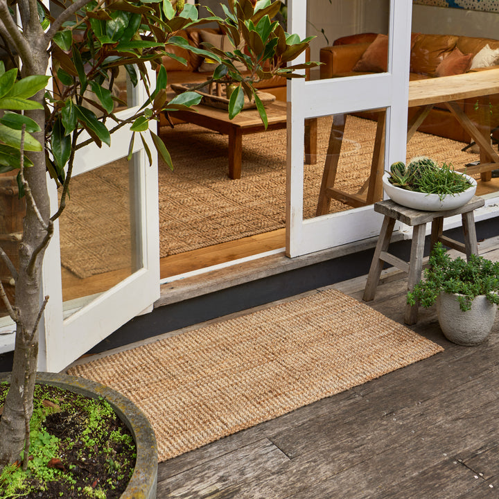 A natural woven jute or sisal doormat leading from a wooden deck onto a patio door into a furnished living room. The scene includes potted green plants and a rustic wooden bench on the patio.