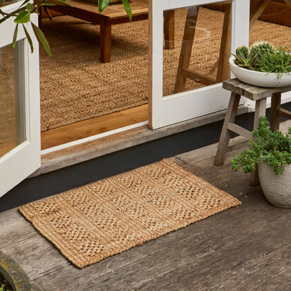 A rectangular natural jute doormat with a textured woven pattern, placed on a weathered wooden deck in front of white-framed glass doors, with potted plants and a small stool nearby.