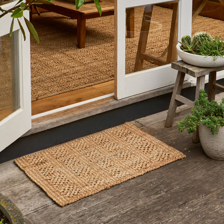 A rectangular natural jute doormat with a textured woven pattern, placed on a weathered wooden deck in front of white-framed glass doors, with potted plants and a small stool nearby.