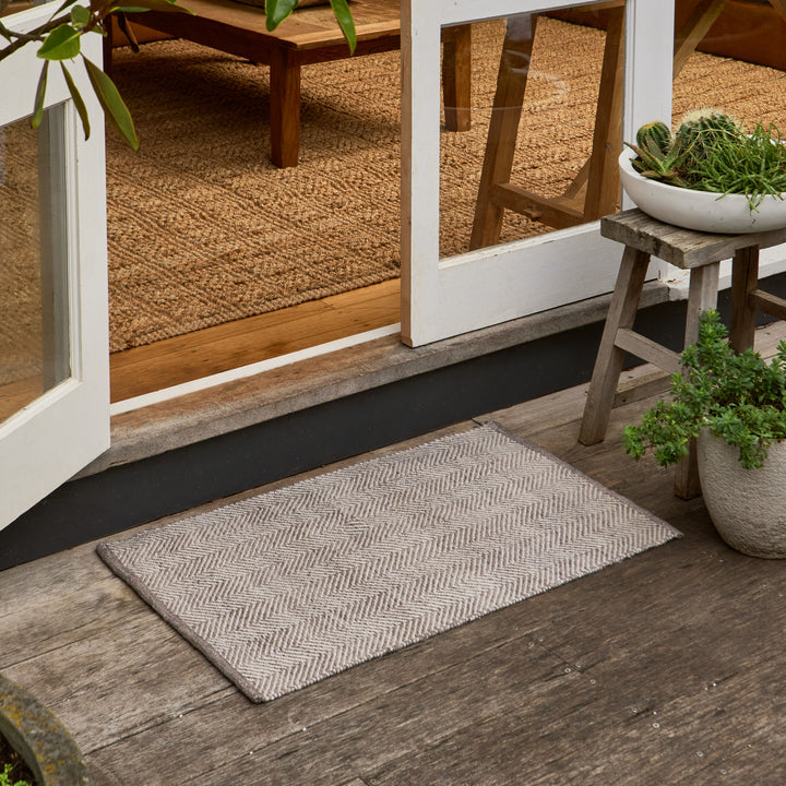 A rectangular woven doormat with a light beige and gray herringbone pattern, placed on a rustic wooden porch in front of white-framed glass doors.