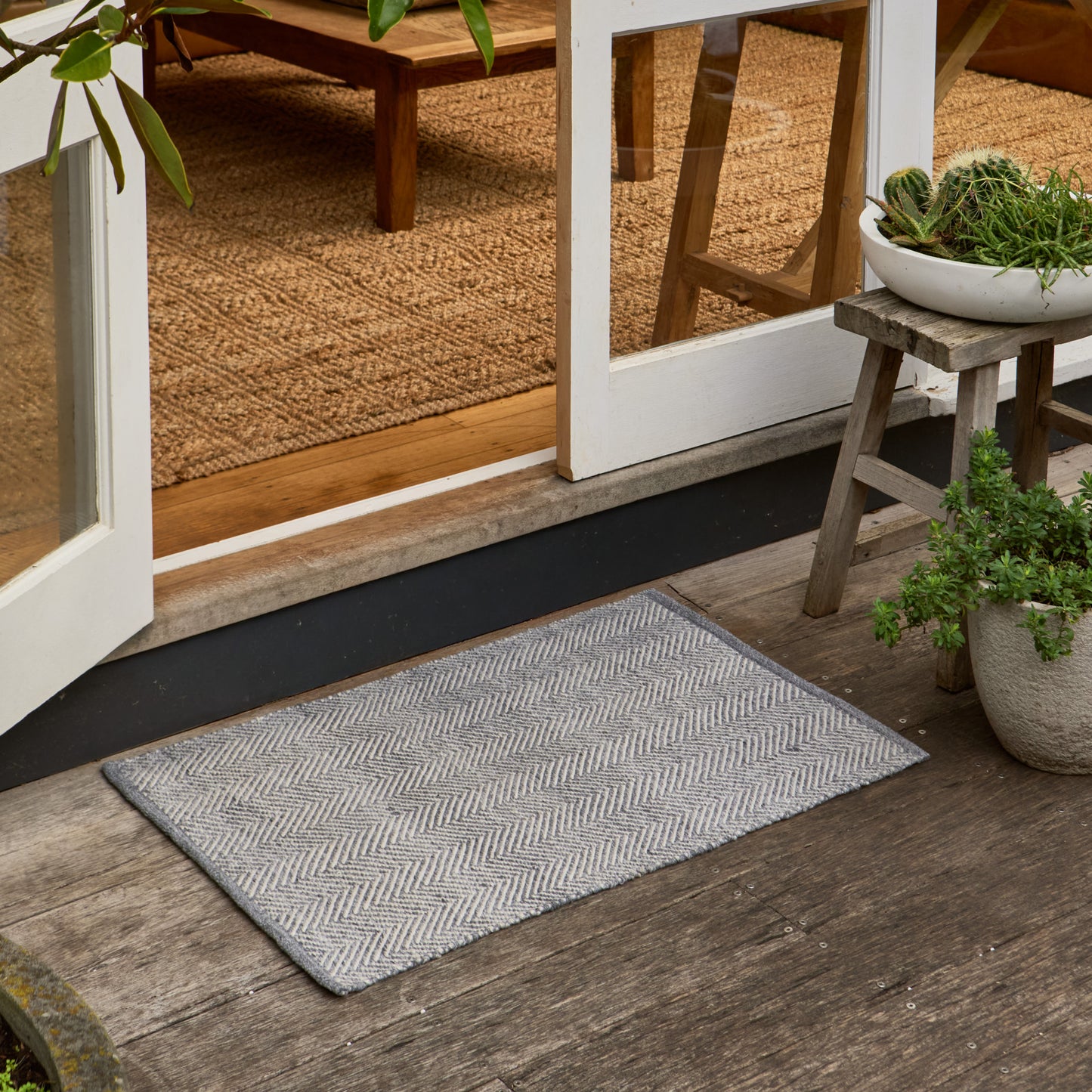 A rectangular gray doormat with a woven herringbone pattern, placed on a weathered wooden deck in front of white-framed glass doors, with potted plants and a stool nearby.
