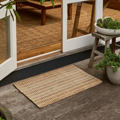 A rectangular natural jute doormat with a woven texture, positioned on a dark wooden porch in front of white-framed glass doors, with potted plants and a small stool on the porch and a woven rug visible inside.