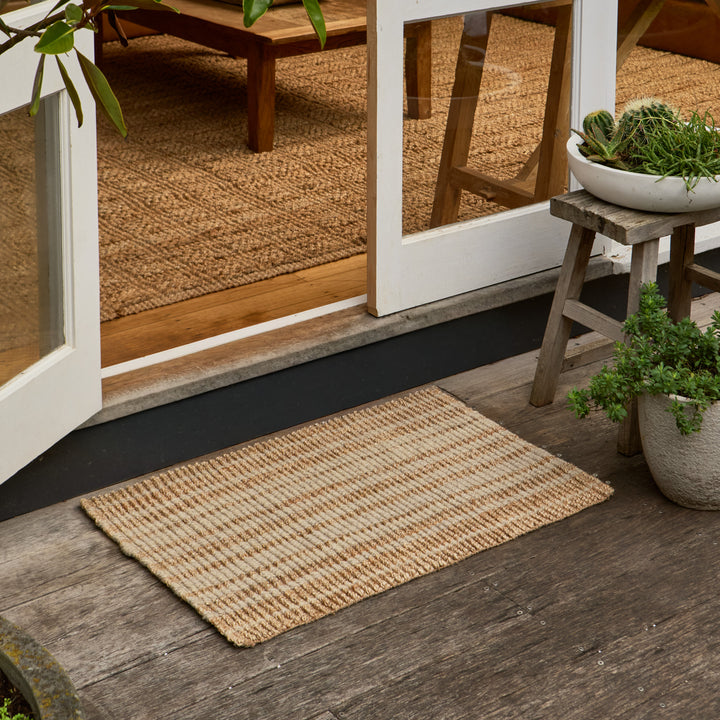 A rectangular natural jute doormat with a woven texture, positioned on a dark wooden porch in front of white-framed glass doors, with potted plants and a small stool on the porch and a woven rug visible inside.