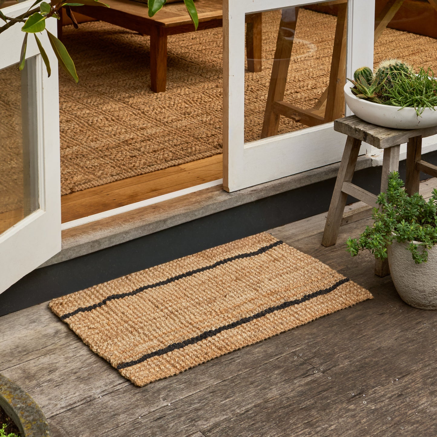 A natural jute doormat with two black stripes, shown on a weathered wooden porch floor in front of glass doors, with potted plants and a stool nearby.