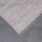 A close-up of a textured area rug with a herringbone pattern in beige and taupe, shown folded over against a gray background.