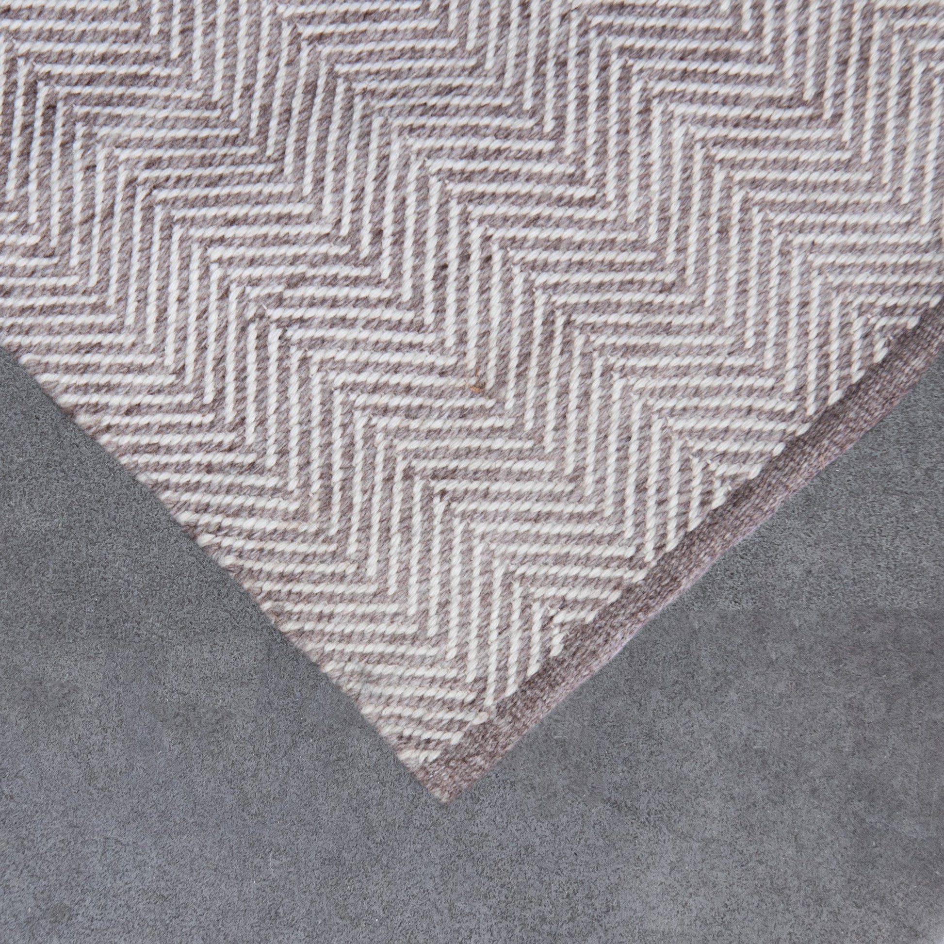 A close-up of a textured area rug with a herringbone pattern in beige and taupe, shown folded over against a gray background.