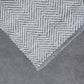 A close-up of a gray and white herringbone patterned rug corner, showing the woven texture and a matching gray border, set against a neutral gray background.