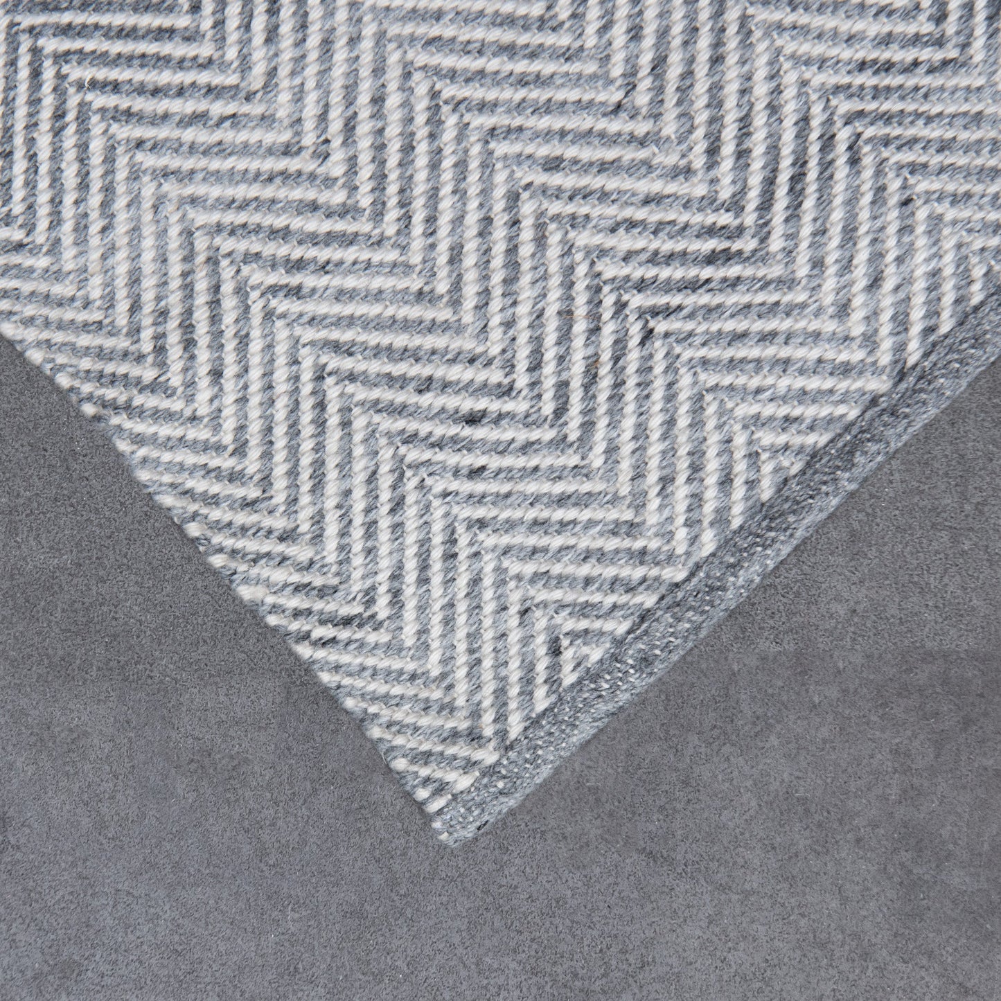 A close-up of a gray and white herringbone patterned rug corner, showing the woven texture and a matching gray border, set against a neutral gray background.